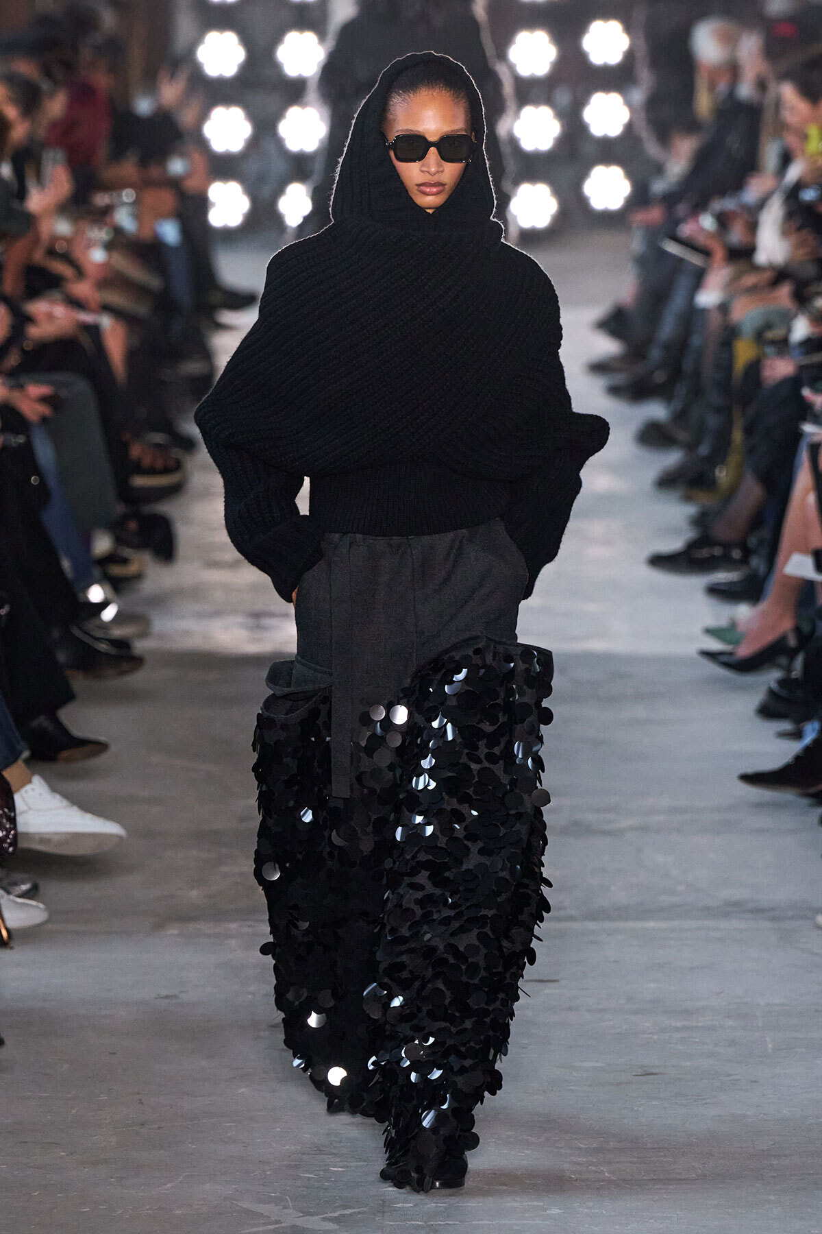 Model walks the runway in a black oversized knit hooded sweater and a floor-length sequined skirt.