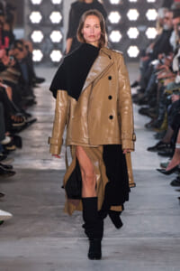 Model walks the runway in a tan leather double‑breasted trench with a black cape draped over one shoulder.