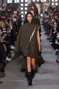 Model walks the runway in an oversized olive-brown layered cape over a matching skirt, with fringed hem and black gloves, audience filming nearby.