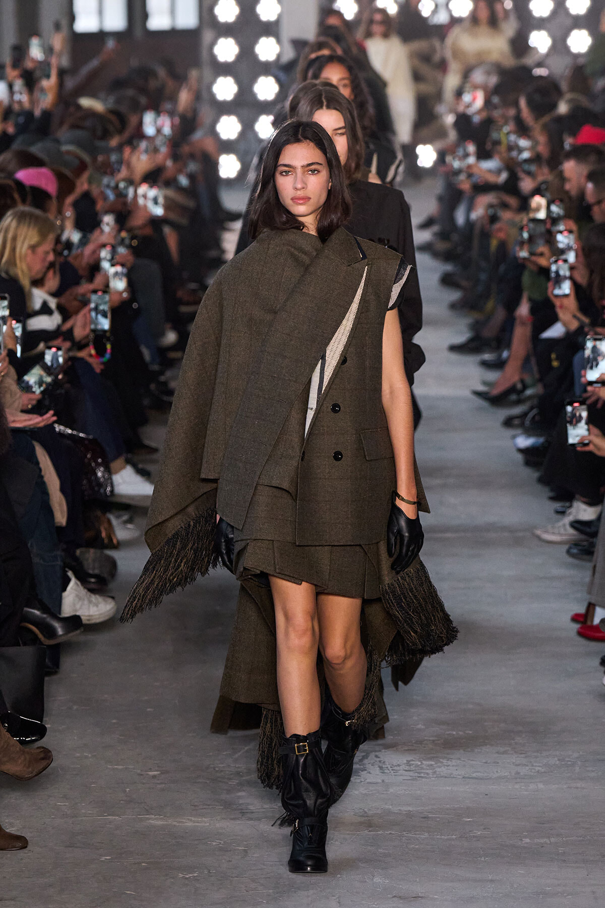Model walks the runway in an oversized olive-brown layered cape over a matching skirt, with fringed hem and black gloves, audience filming nearby.