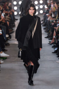 Model walks a fashion show wearing an all-black layered outfit with a fringed cape and oversized coat, carrying a small black bag and knee-high boots.
