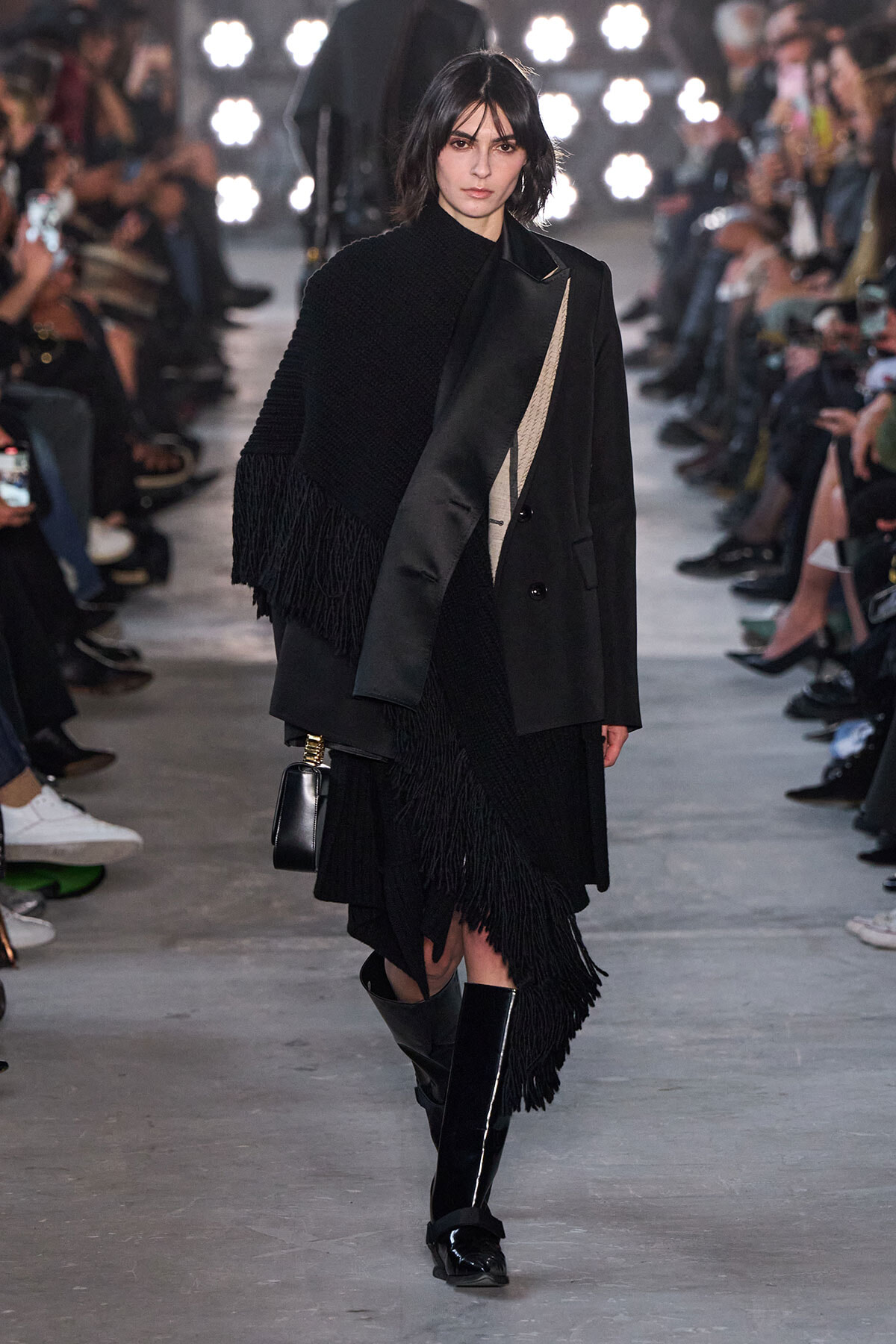 Model walks a fashion show wearing an all-black layered outfit with a fringed cape and oversized coat, carrying a small black bag and knee-high boots.