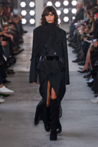 Model walks a fashion show in an all-black, layered coat with a wide belt and high slit, wearing knee-high boots.