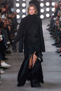 Model on a runway wearing an oversized black blazer with draped sheer scarf and long layered skirt, walking toward camera.