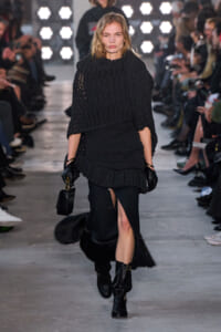 Model walks a fashion runway in an all-black layered outfit: oversized knit poncho, fitted skirt with fur trim, and knee-high black boots, carrying a small black handbag and wearing gloves.