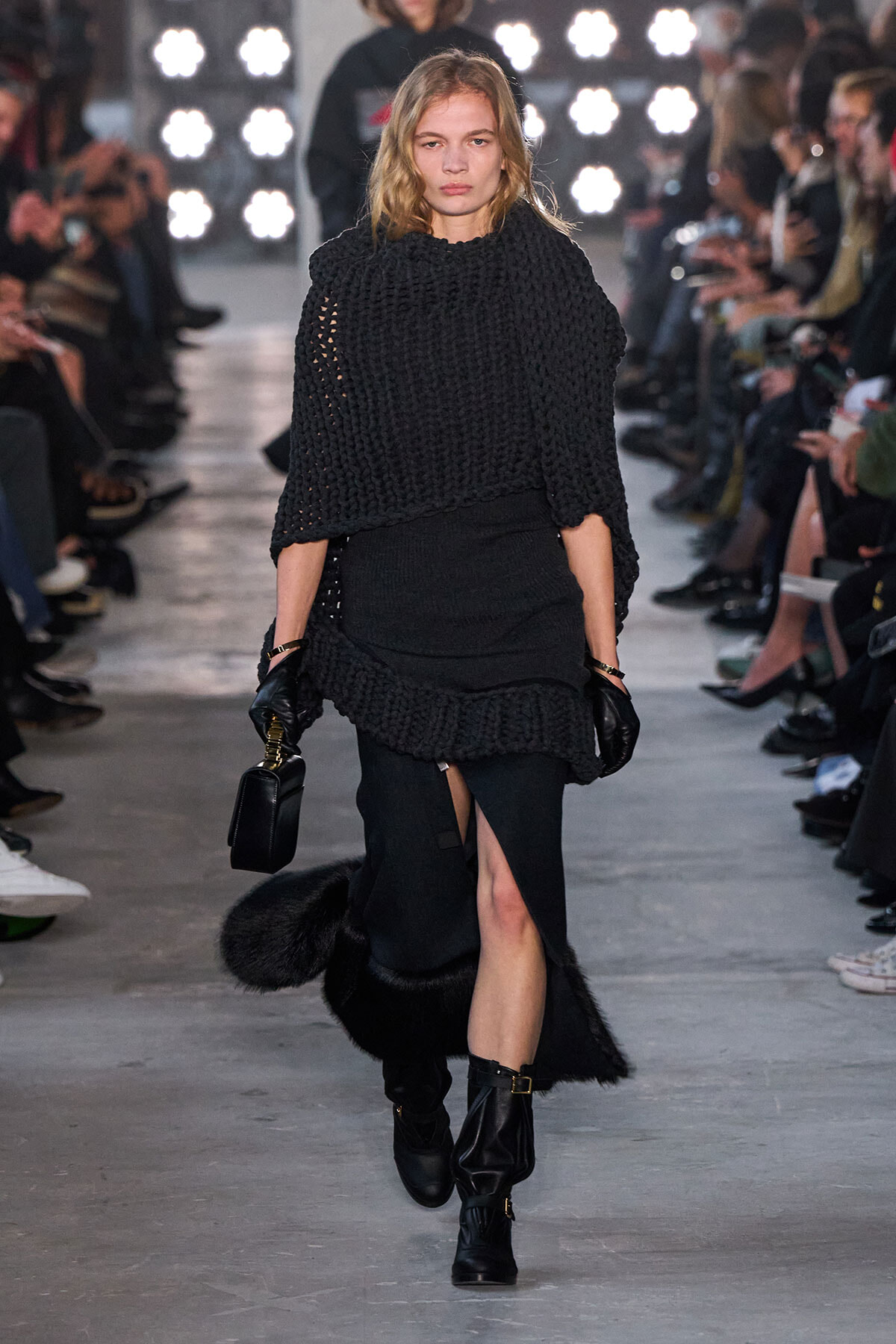 Model walks a fashion runway in an all-black layered outfit: oversized knit poncho, fitted skirt with fur trim, and knee-high black boots, carrying a small black handbag and wearing gloves.