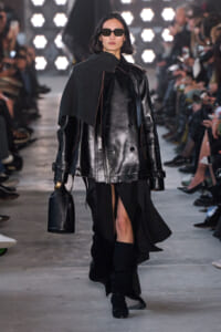 Model walks the runway in a black leather oversized coat with a gray cape draped over one shoulder, wearing sunglasses and carrying a black handbag.