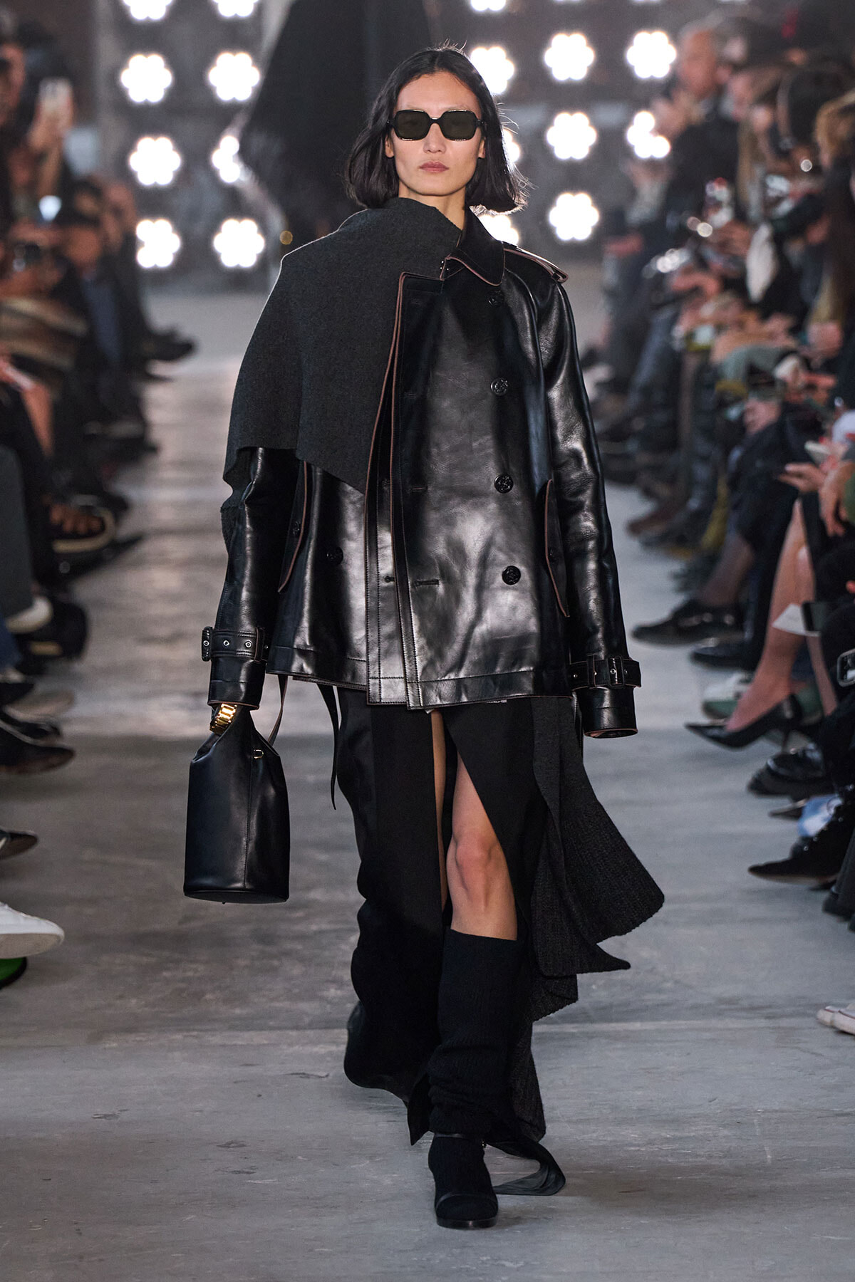 Model walks the runway in a black leather oversized coat with a gray cape draped over one shoulder, wearing sunglasses and carrying a black handbag.