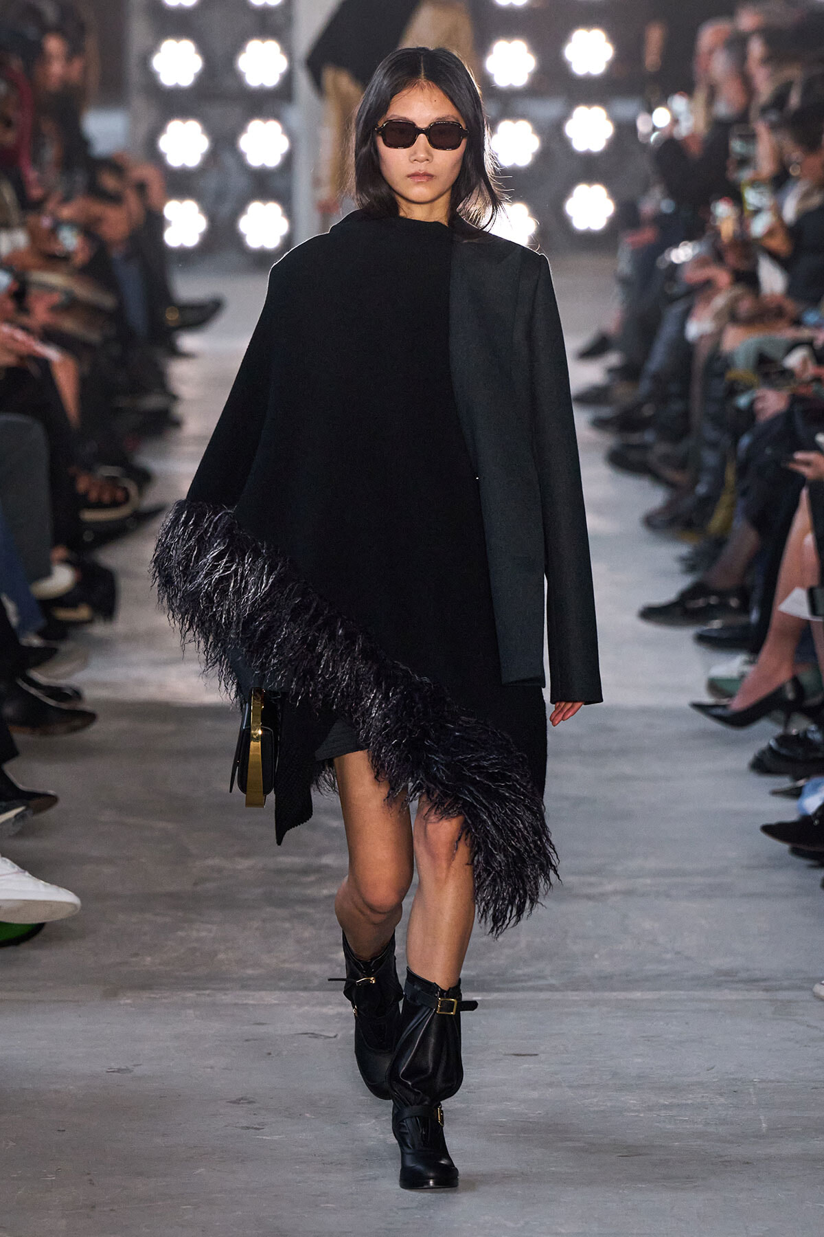 Model walks runway in black outfit with fringe at hem, wearing dark sunglasses and ankle boots.
