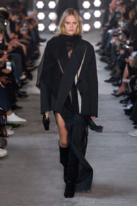 Model walking a runway in a black layered outfit with a sheer top, oversized blazer, and flowing fabric; audience on both sides.