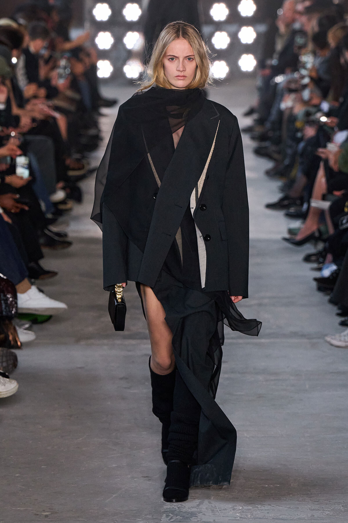 Model walking a runway in a black layered outfit with a sheer top, oversized blazer, and flowing fabric; audience on both sides.