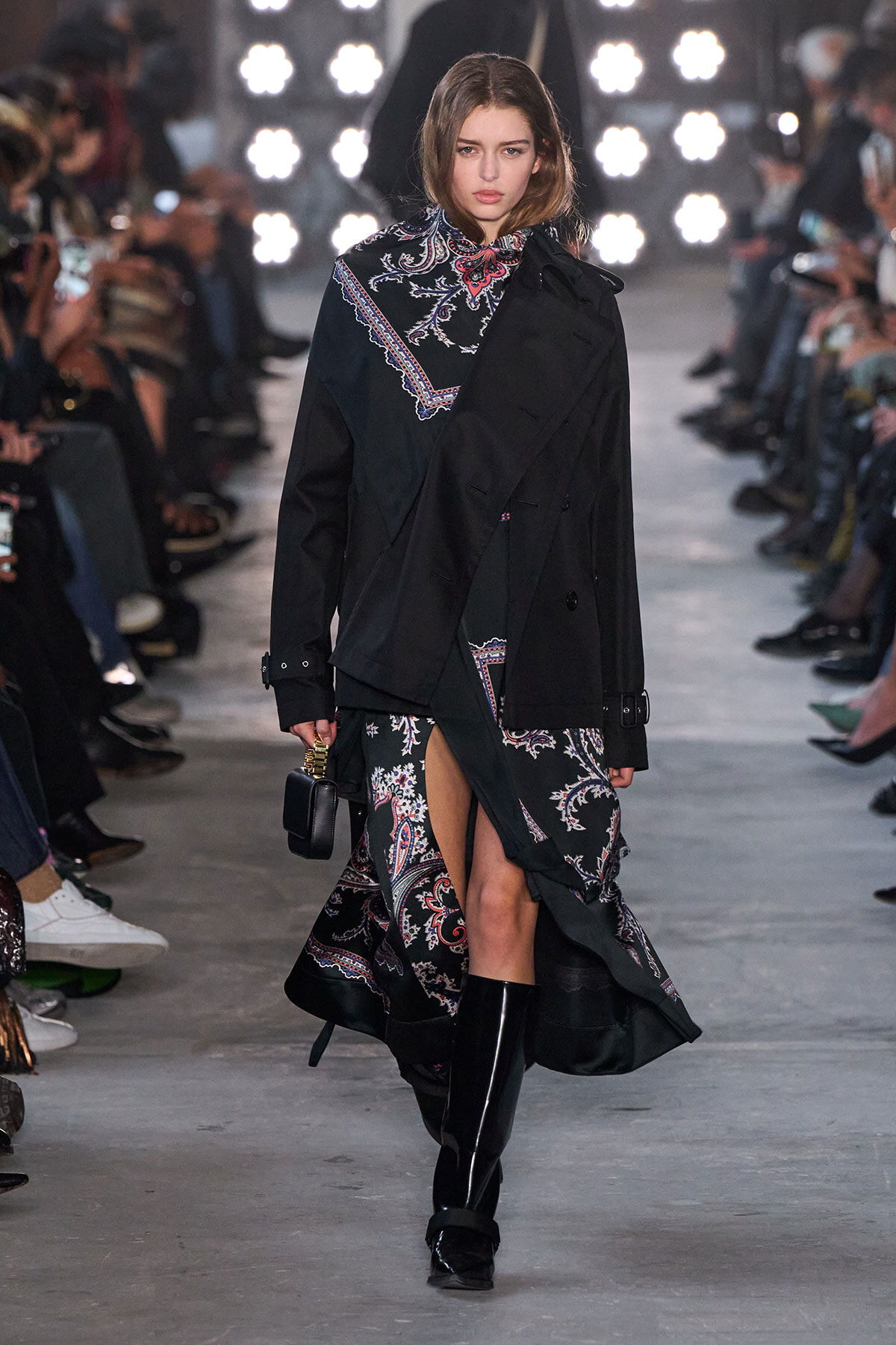 Runway model walks wearing a black oversized trench over a paisley-patterned dress with a high slit and shiny black knee-high boots.