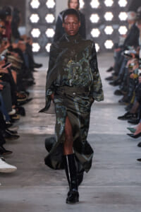 Model on a runway wearing a dark green paisley-print blouse with a matching skirt and knee-high black boots.
