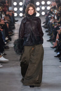 Model on a runway wearing a dark leather jacket with a furry collar and a long olive skirt, audience visible in the background.