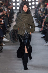 Model walking a fashion show in an oversized olive parka, black leather skirt with a front slit, and black boots, carrying a furry bag.