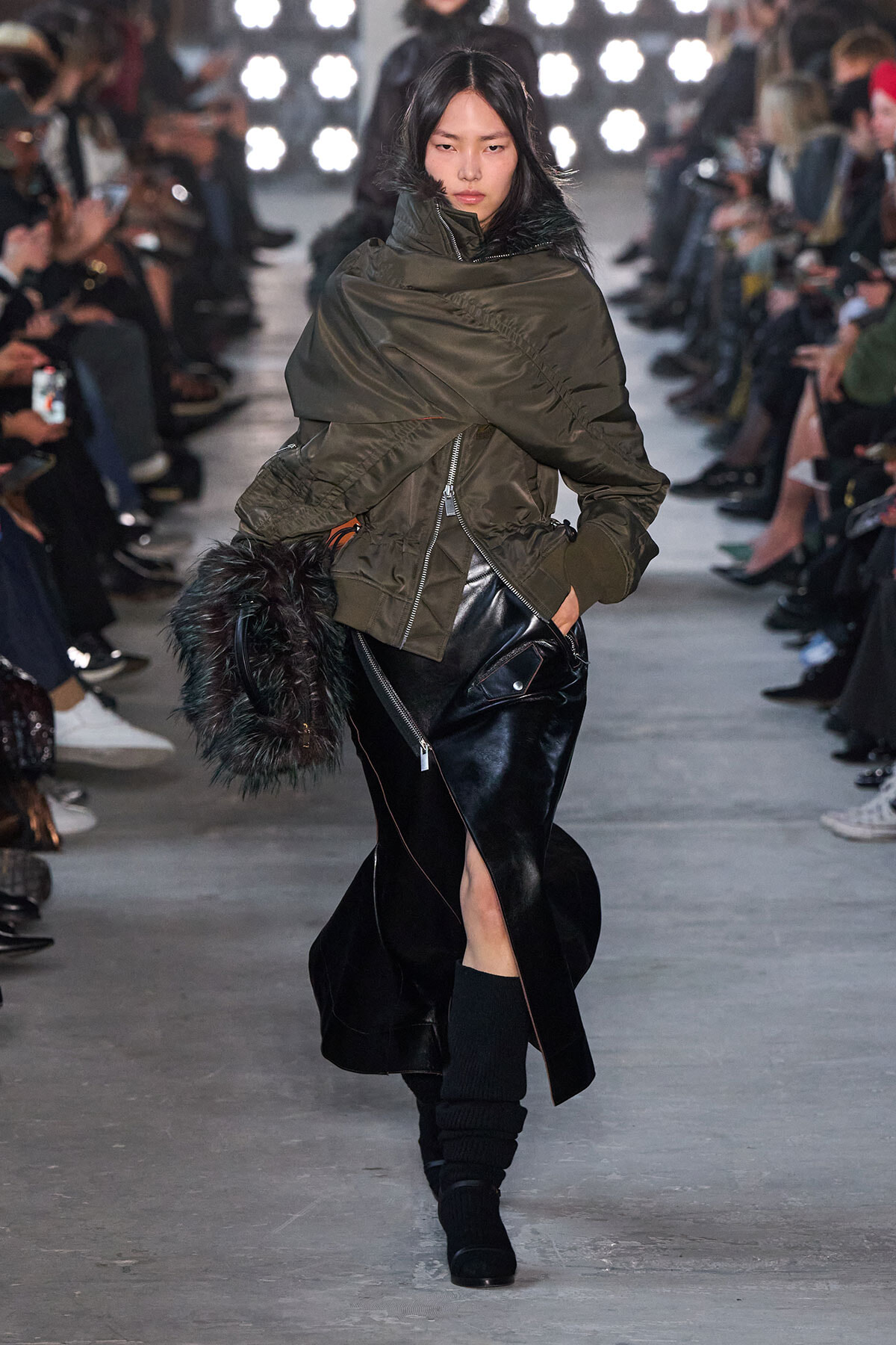 Model walking a fashion show in an oversized olive parka, black leather skirt with a front slit, and black boots, carrying a furry bag.