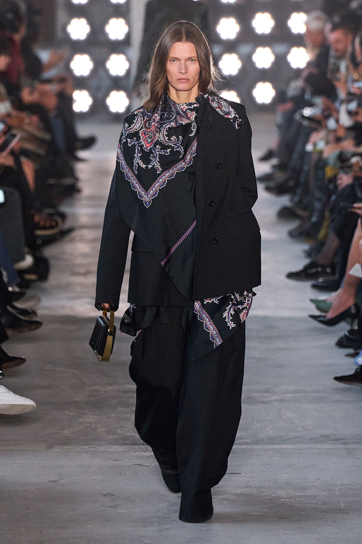 Model walks the runway in a black suit with a paisley-patterned scarf draped over the shoulder, audience lit by bright backstage lights in the background.