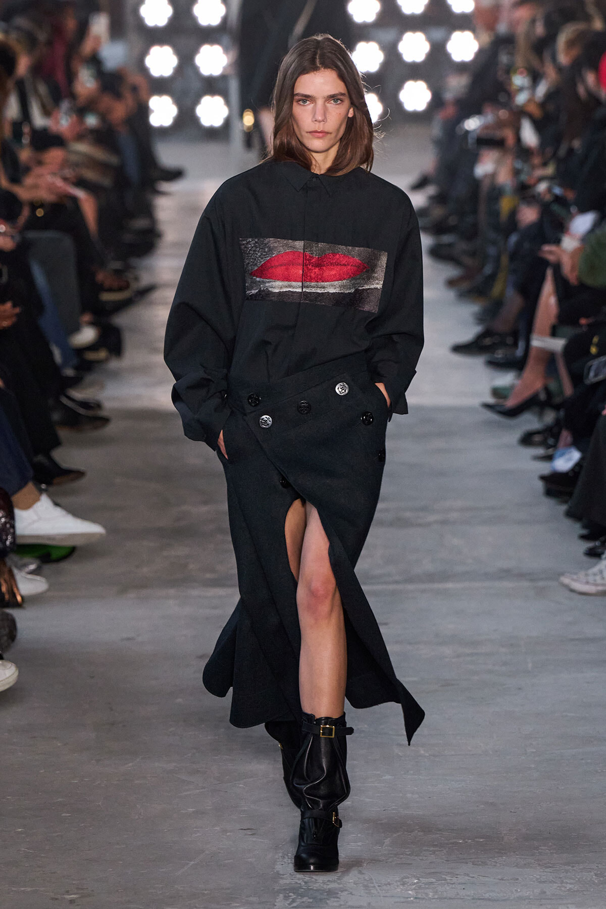 Model walking the runway in a black oversized shirt with a red lips graphic, paired with a high-slit black skirt and black ankle boots.