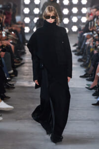 Model walking the runway in an all-black layered outfit: oversized textured turtleneck, long coat, and wide-leg pants, paired with dark sunglasses and blonde hair. Audience sits along the catwalk with bright stage lights in the background.