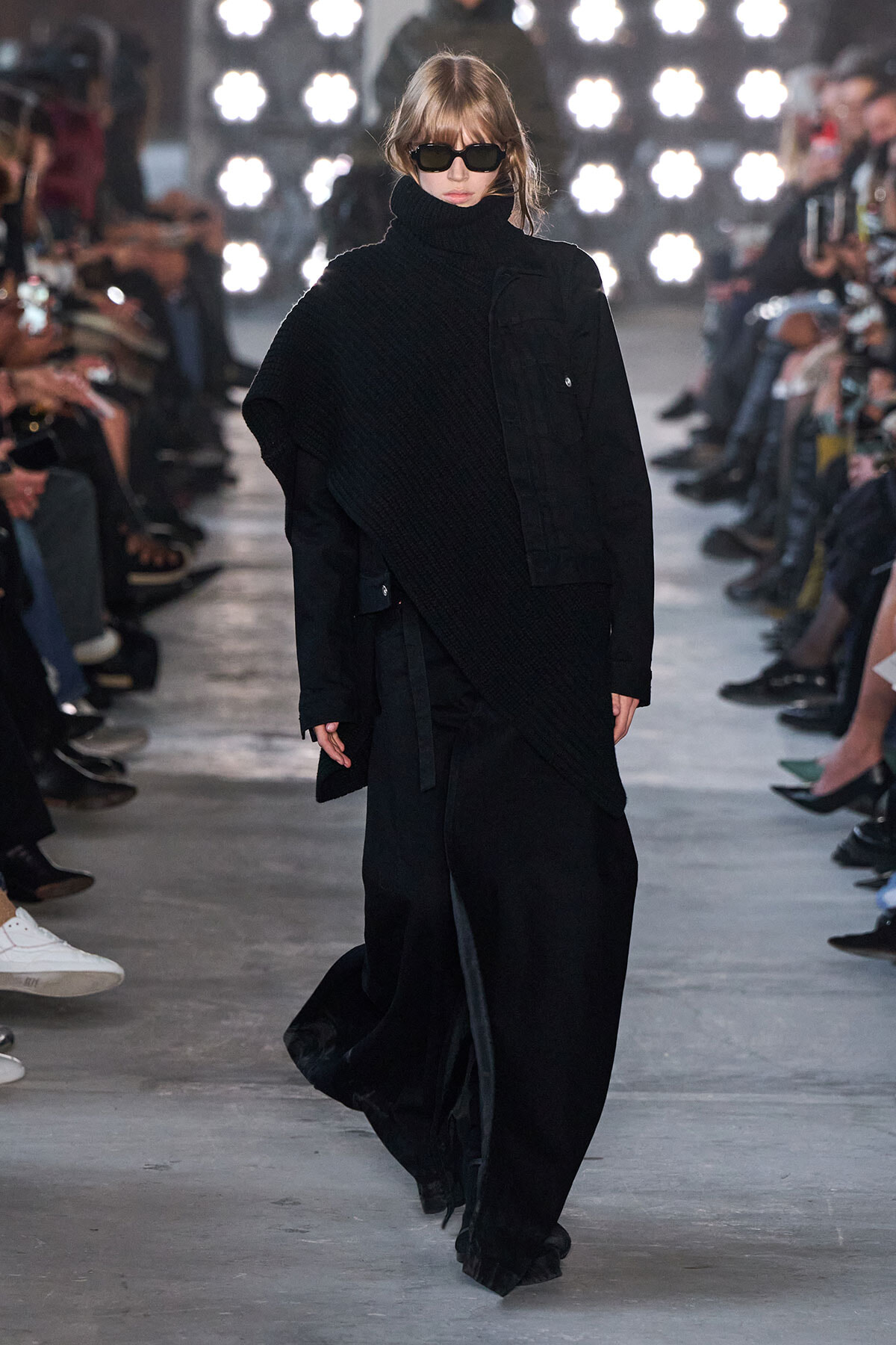 Model walking the runway in an all-black layered outfit: oversized textured turtleneck, long coat, and wide-leg pants, paired with dark sunglasses and blonde hair. Audience sits along the catwalk with bright stage lights in the background.