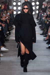 Model walks a fashion show wearing a black oversized cloak-like coat, knee-high black boots and sunglasses on a lit runway.
