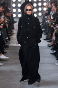 Model in an all-black oversized utility jacket and wide-legged pants walks the runway, wearing sunglasses with a stern expression.