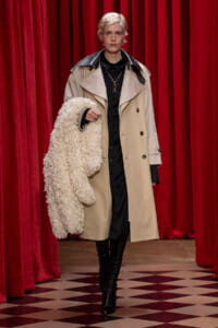 Runway model in a beige trench over black clothing, holding a fluffy cream coat, with red velvet curtains in the background.