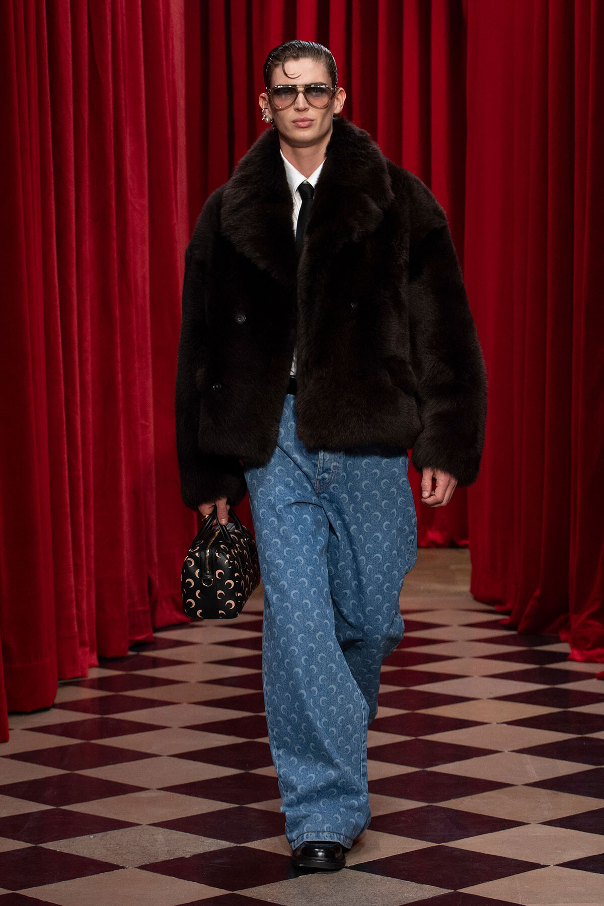 Model in a black fur coat over a white shirt and black tie, with blue patterned wide-leg jeans, oversized sunglasses, and a crescent-pattern handbag against red curtains.
