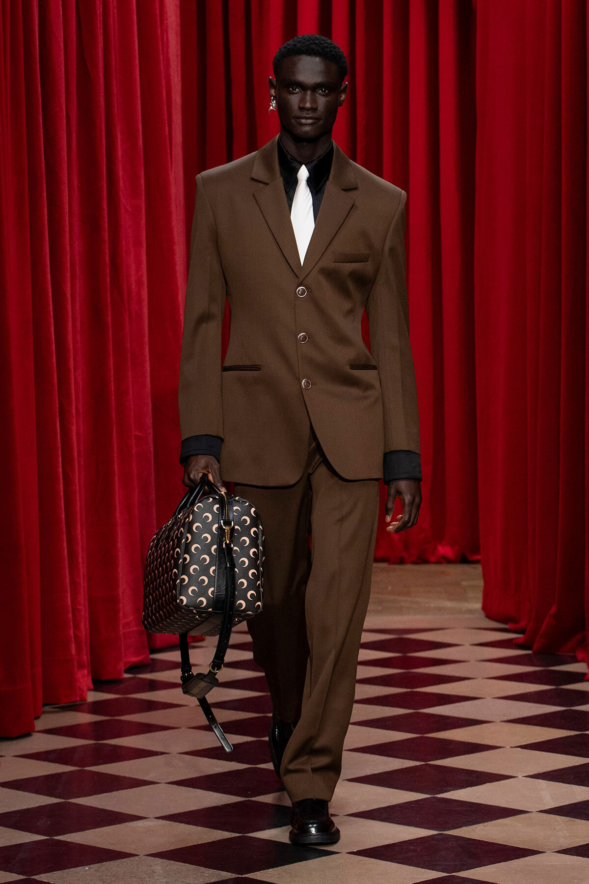 Male fashion model in a brown suit with a white tie walks the runway, carrying a patterned handbag against red curtains.