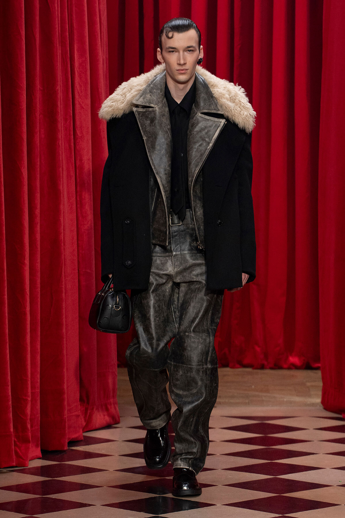 Male model on a runway wearing a black coat with a cream shearling collar, gray distressed pants, black shirt, and carrying a small black handbag against red velvet curtains.