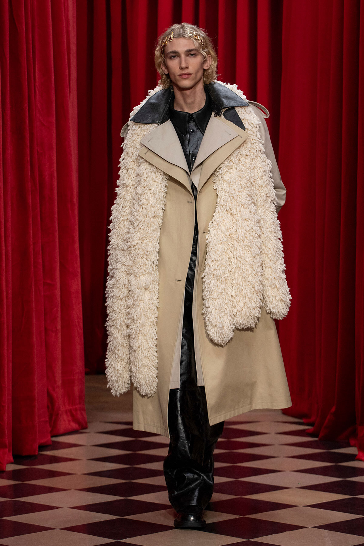 Model on a runway wearing a beige trench with a chunky cream shearling scarf, black leather outfit, red velvet backdrop