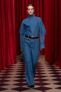 Model wearing a blue denim jumpsuit with an oversized sleeves and black belt, walking on a checkered floor in front of red velvet curtains.