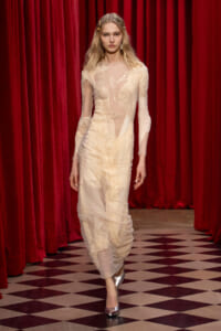 Female model walks the runway in a sheer cream lace gown with red curtains in the background.