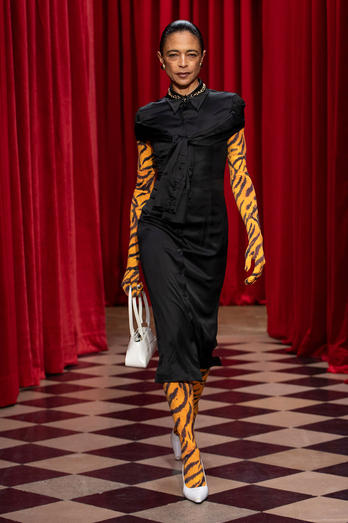 Model walking a runway in a black midi dress, orange tiger-print gloves and stockings, white heels, and a white handbag against red curtains.
