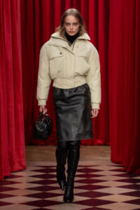 Model on a runway wearing a cream puffer jacket, black leather skirt, knee-high boots, and a small black handbag against red velvet curtains.