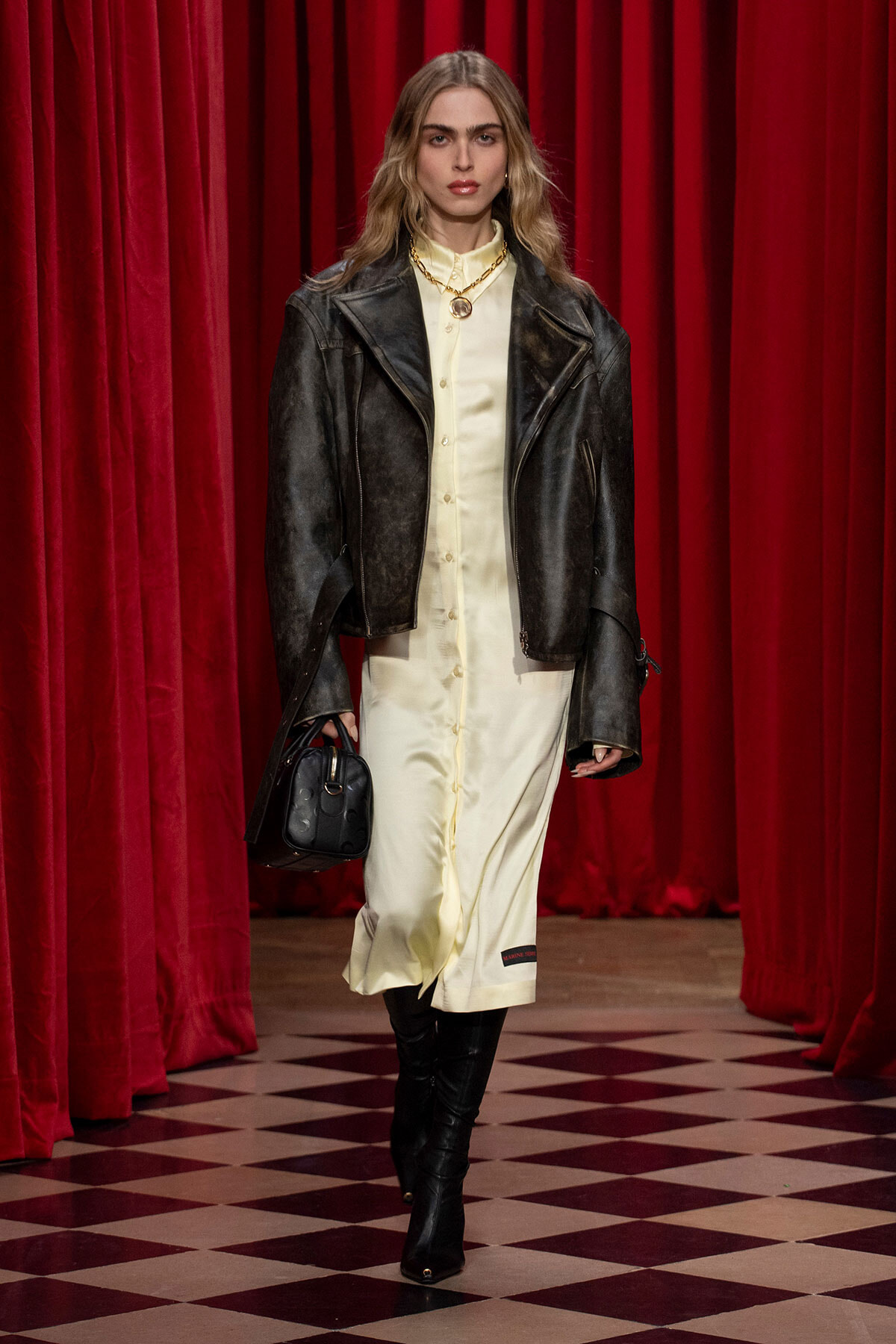 Model on a runway wearing a cream shirt dress with a black leather jacket, gold necklace, black knee-high boots, and a black handbag against red velvet curtains.