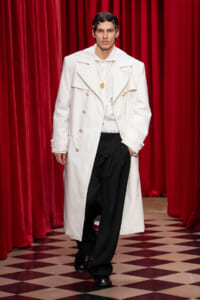 Model wears a long white trench coat over a white shirt and black pants, walking on a checkered floor with red stage curtains behind them.