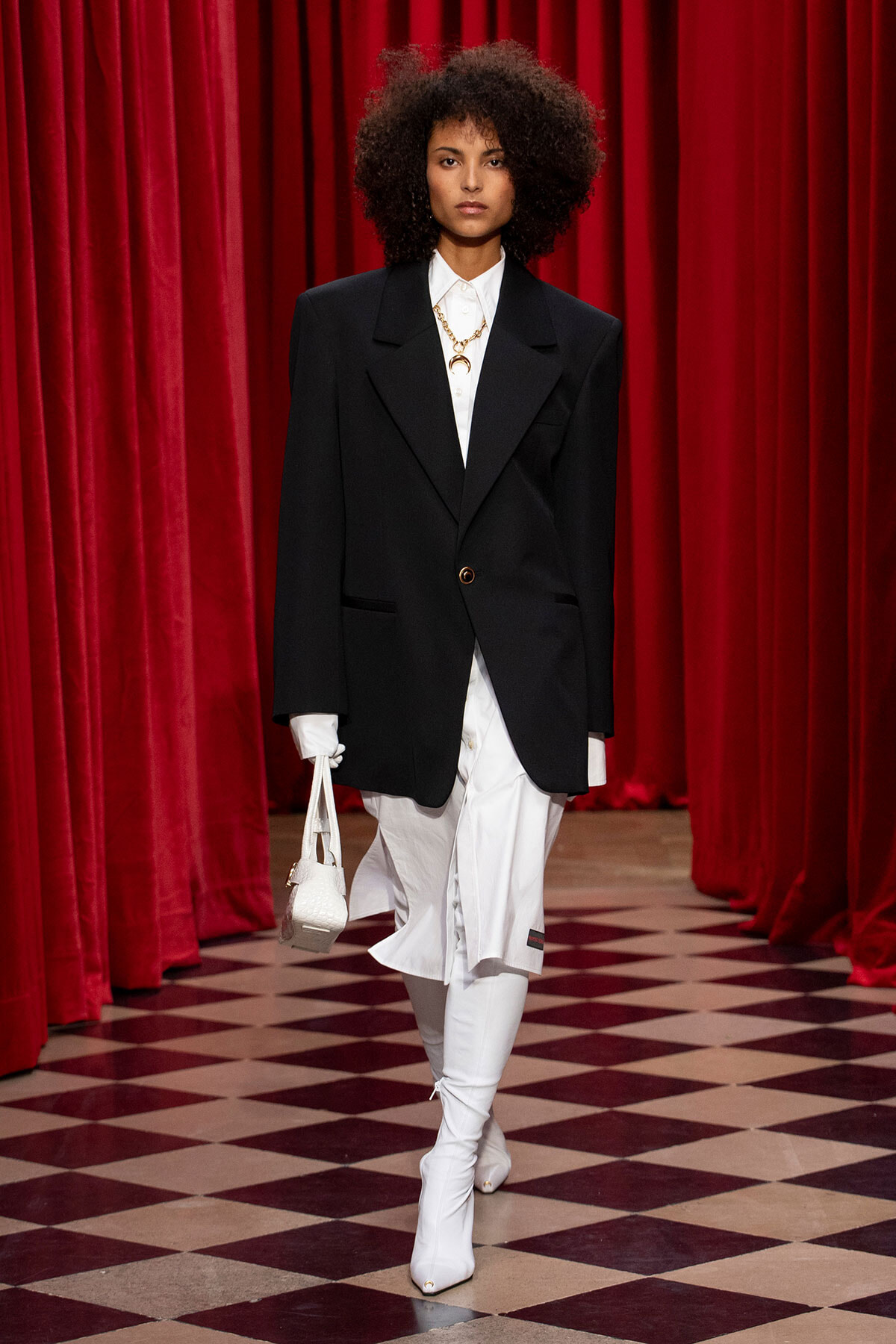 Model on runway wearing an oversized black blazer over a white shirt and pants, gold necklace, white knee-high boots, and a white handbag in front of red curtains.