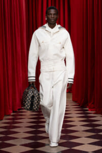 Model walks a runway in a white utility jumpsuit with striped collar and cuffs, carrying a patterned black bag against red curtains.