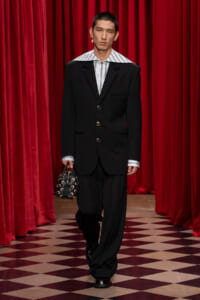 Male model on a runway wearing an oversized black suit with a striped shirt, carrying a small patterned handbag against red velvet curtains.