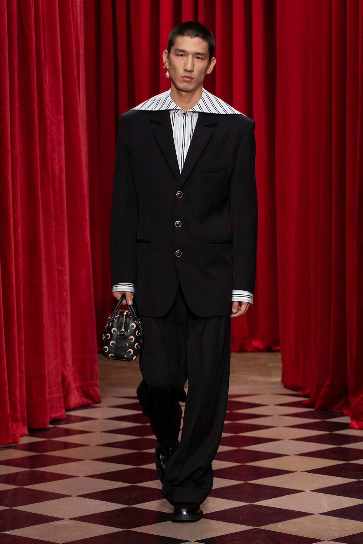 Male model on a runway wearing an oversized black suit with a striped shirt, carrying a small patterned handbag against red velvet curtains.