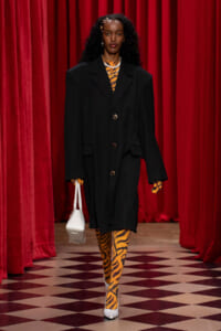 Runway model in an oversized black coat over tiger-print clothing, carrying a white handbag against red velvet curtains and a checkered floor.
