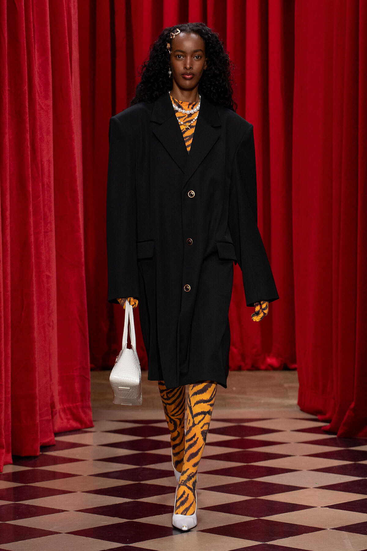 Runway model in an oversized black coat over tiger-print clothing, carrying a white handbag against red velvet curtains and a checkered floor.