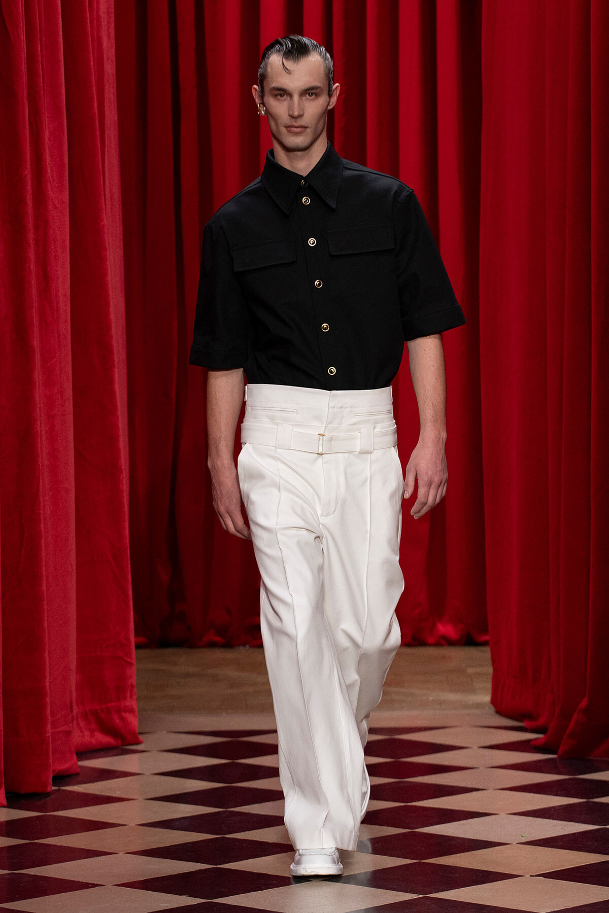 Male model walks a runway wearing a black short-sleeve button-up shirt with gold buttons and white high-waisted trousers against red velvet curtains.