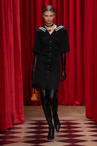 Runway model in a black button-down dress with a white-striped sailor collar, black gloves, knee-high boots, and a small tan handbag against red theater curtains.