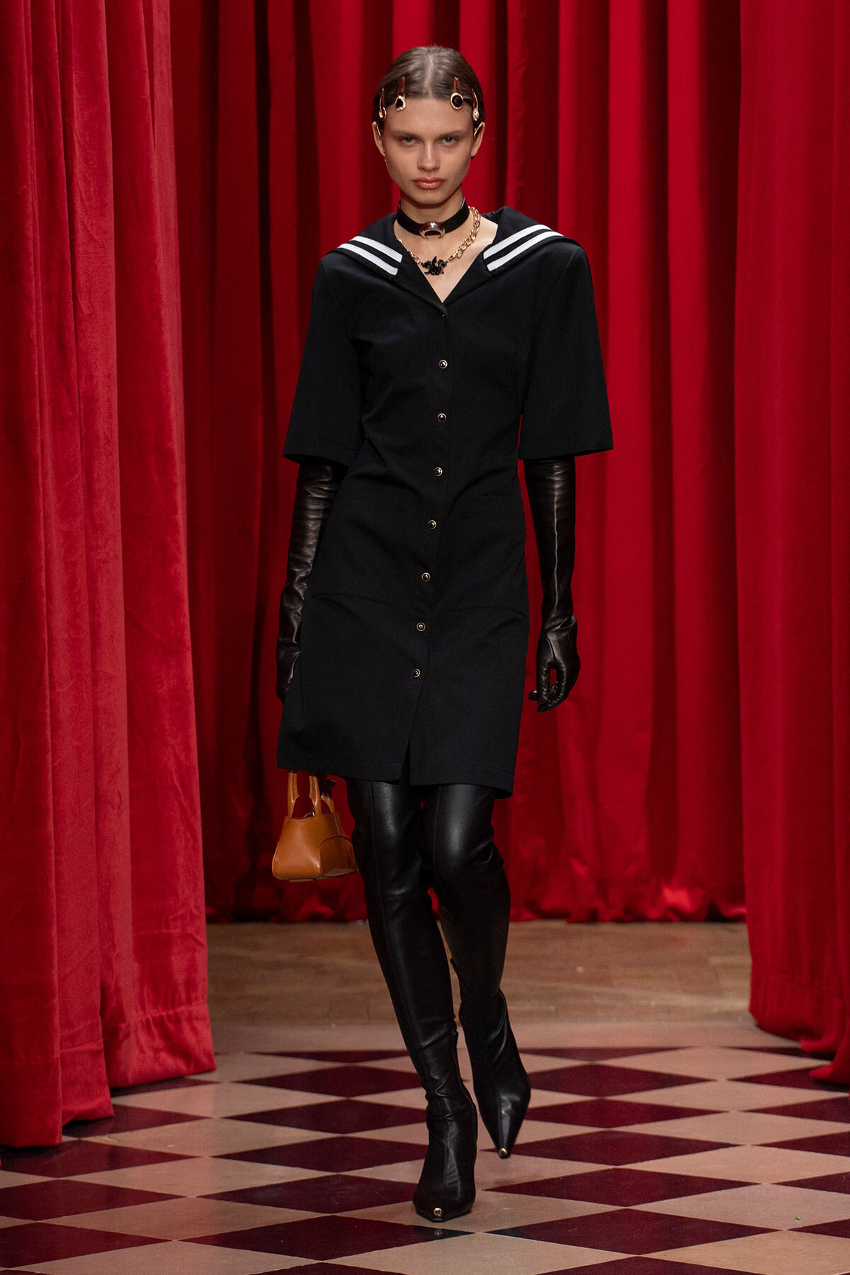 Runway model in a black button-down dress with a white-striped sailor collar, black gloves, knee-high boots, and a small tan handbag against red theater curtains.