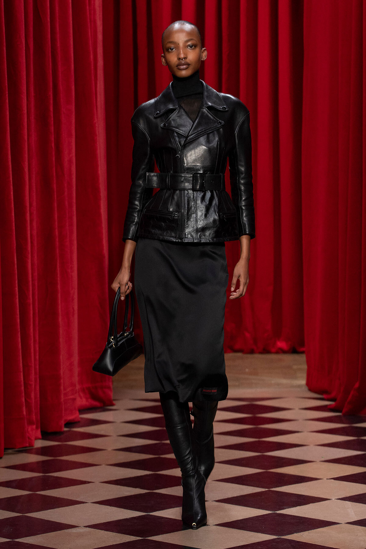 Model wearing a black leather belted jacket, black turtleneck, and a black midi skirt with knee-high boots on a red-curtained runway.