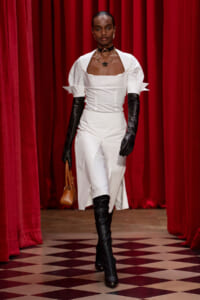 Model walks the runway in a white square-neck dress with puff sleeves, black opera gloves, knee-high boots, and a small tan handbag.