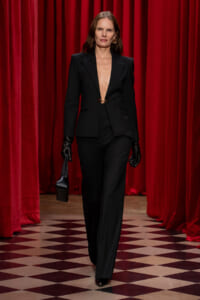 Woman in a black tailored suit with a deep plunging neckline walks on a checkered floor, red velvet curtains behind her.