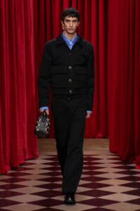 Male model walks a runway in a black utility jacket and matching trousers, blue shirt, and a small patterned handbag, against red velvet curtains and a checkered floor backdrop.